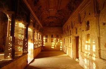 An ancient architectural space features intricately carved walls and ceilings. Sunlight streams through detailed latticework, casting patterned shadows on the floor and walls. The interior is beautifully weathered, with an earthy color palette dominated by browns and golds.