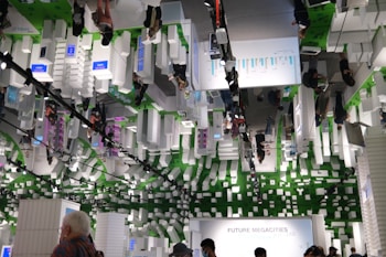 An indoor exhibition space with a futuristic theme featuring white, block-like structures and several people moving through the area. The ceiling mirrors the scene below, adding to the visual complexity. Green elements and digital display panels are integrated into the design. The space has an informational focus, likely about urban development or technology.