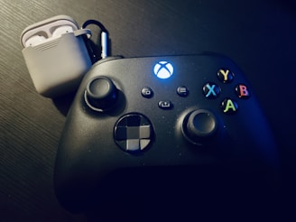 A black gaming controller with illuminated buttons sits on a dark surface. Nearby, a pair of wireless earbuds in a white charging case is visible.