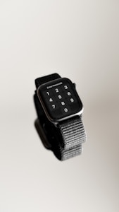 A sleek smartwatch displays a passcode entry screen featuring digits from 0 to 9. The device has a dark screen surrounded by a metallic frame and is attached to a textured black strap. The background is a smooth, neutral tone, creating a minimalist aesthetic.