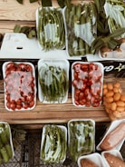 A variety of fresh vegetables and fruits are arranged in white plastic trays on a wooden surface. The produce includes green peppers, tomatoes, cucumbers, and possibly oranges or kumquats. Each tray is wrapped in clear plastic, and there are leaves placed around some of the trays, adding a rustic touch to the display.
