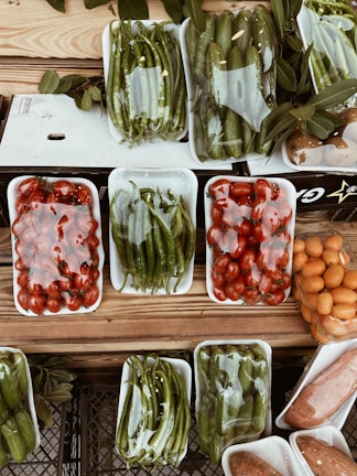 A variety of fresh vegetables and fruits are arranged in white plastic trays on a wooden surface. The produce includes green peppers, tomatoes, cucumbers, and possibly oranges or kumquats. Each tray is wrapped in clear plastic, and there are leaves placed around some of the trays, adding a rustic touch to the display.