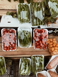 A variety of fresh vegetables and fruits are arranged in white plastic trays on a wooden surface. The produce includes green peppers, tomatoes, cucumbers, and possibly oranges or kumquats. Each tray is wrapped in clear plastic, and there are leaves placed around some of the trays, adding a rustic touch to the display.