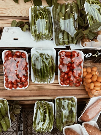 A variety of fresh vegetables and fruits are arranged in white plastic trays on a wooden surface. The produce includes green peppers, tomatoes, cucumbers, and possibly oranges or kumquats. Each tray is wrapped in clear plastic, and there are leaves placed around some of the trays, adding a rustic touch to the display.