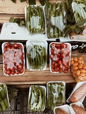 A variety of fresh vegetables and fruits are arranged in white plastic trays on a wooden surface. The produce includes green peppers, tomatoes, cucumbers, and possibly oranges or kumquats. Each tray is wrapped in clear plastic, and there are leaves placed around some of the trays, adding a rustic touch to the display.