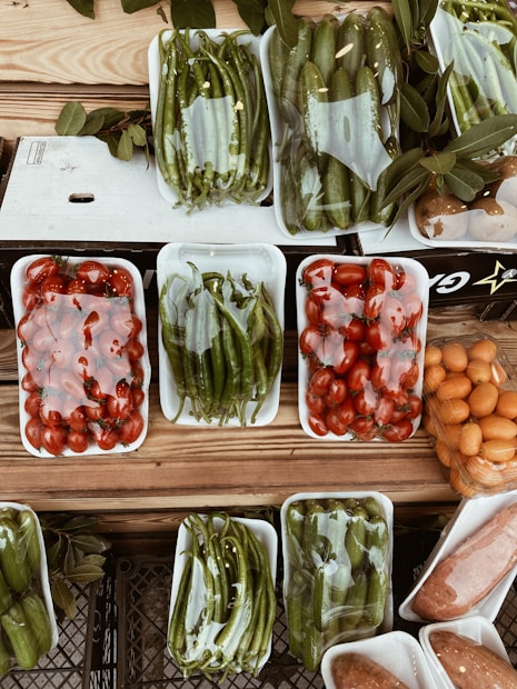 A variety of fresh vegetables and fruits are arranged in white plastic trays on a wooden surface. The produce includes green peppers, tomatoes, cucumbers, and possibly oranges or kumquats. Each tray is wrapped in clear plastic, and there are leaves placed around some of the trays, adding a rustic touch to the display.