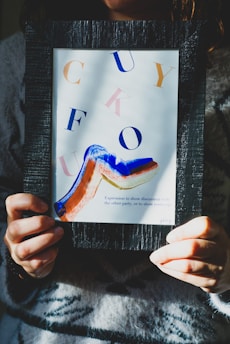 A person holds a framed piece of abstract art that features scattered letters and a bold, colorful paint stroke. The letters are in various colors such as orange, blue, and pink. The frame is black and textured, while the artwork contains a mix of blue, orange, and red colors in its brush stroke. The person is wearing a patterned sweater.