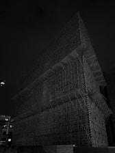 A large, multi-layered scaffolding structure stands prominently against a dark night sky. The intricate network of metal bars and platforms is illuminated by minimal lighting, creating a stark, geometric pattern. A surveillance camera is visible in the lower left corner, observing the construction site.