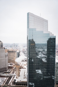 A high-rise building with a modern glass facade reflecting the urban landscape of a city. The surrounding area contains a mix of other skyscrapers and smaller buildings, with visible streets below. The sky is overcast, giving the scene a cold, subdued appearance.