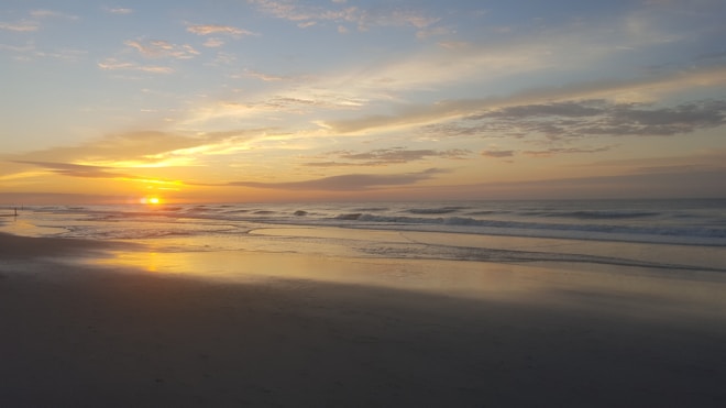 A serene beach scene with a vibrant sunrise casting warm hues across the sky and water. Gentle waves wash onto the shore, and the expansive sky is partially covered with scattered, wispy clouds.