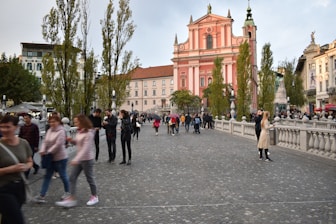 A lively cityscape featuring a large, ornate pink church with detailed architecture, surrounded by lush greenery from tall trees. The area is bustling with numerous people walking along a wide cobblestone pathway, some stopping to take pictures or chat. The buildings lining the street exhibit varied architectural styles, enhancing the urban charm.