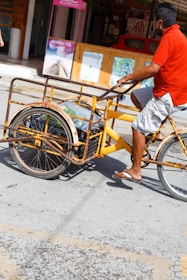 A person wearing a red shirt and gray shorts is riding a yellow tricycle cart on a sunny street. The cart is rusted with large wheels and appears to be used for carrying items. In the background, there are shop signs and advertisements, including one for Chichen Itza.