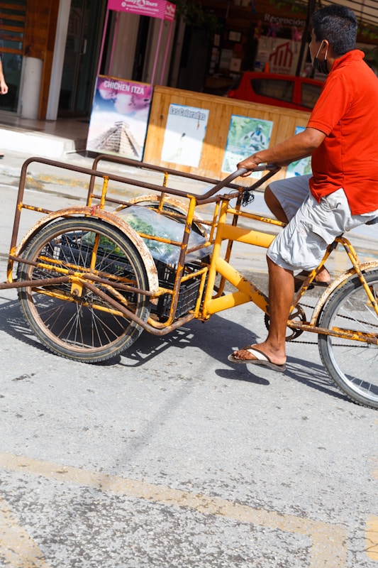 A person wearing a red shirt and gray shorts is riding a yellow tricycle cart on a sunny street. The cart is rusted with large wheels and appears to be used for carrying items. In the background, there are shop signs and advertisements, including one for Chichen Itza.