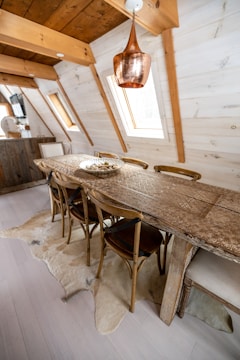 A rustic dining area features a long wooden table with intricate carvings, surrounded by wooden chairs in an attic-style room. The space is adorned with a large copper pendant light hanging from the ceiling. The walls and ceiling are paneled with light-colored wood, and natural light streams in through skylight windows. A beige rug with a fur-like texture is placed underneath the table.