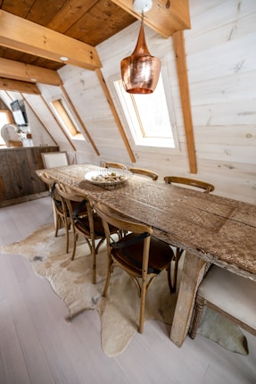 A rustic dining area features a long wooden table with intricate carvings, surrounded by wooden chairs in an attic-style room. The space is adorned with a large copper pendant light hanging from the ceiling. The walls and ceiling are paneled with light-colored wood, and natural light streams in through skylight windows. A beige rug with a fur-like texture is placed underneath the table.