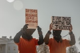 Two individuals dressed in orange jumpsuits and hoods are holding protest signs. One sign reads '20 Years of Disgrace Close Guantanamo' and the other says 'I Am Dying Waiting for Justice'. They are chained and standing outside a significant government building.