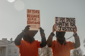 Two individuals dressed in orange jumpsuits and hoods are holding protest signs. One sign reads '20 Years of Disgrace Close Guantanamo' and the other says 'I Am Dying Waiting for Justice'. They are chained and standing outside a significant government building.