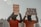 Two individuals dressed in orange jumpsuits and hoods are holding protest signs. One sign reads '20 Years of Disgrace Close Guantanamo' and the other says 'I Am Dying Waiting for Justice'. They are chained and standing outside a significant government building.