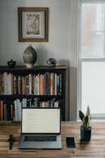A home office setup features a wooden desk with a laptop, smartphone, and a smartwatch placed on it. Behind the desk, there is a bookshelf filled with a variety of books, and atop the bookshelf are decorative items including a large vase and a bowl. A framed artwork hangs above the bookshelf. To the right, there is a window letting in natural light, and a small potted plant adds a touch of greenery to the scene.