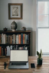 A home office setup features a wooden desk with a laptop, smartphone, and a smartwatch placed on it. Behind the desk, there is a bookshelf filled with a variety of books, and atop the bookshelf are decorative items including a large vase and a bowl. A framed artwork hangs above the bookshelf. To the right, there is a window letting in natural light, and a small potted plant adds a touch of greenery to the scene.