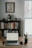 A home office setup features a wooden desk with a laptop, smartphone, and a smartwatch placed on it. Behind the desk, there is a bookshelf filled with a variety of books, and atop the bookshelf are decorative items including a large vase and a bowl. A framed artwork hangs above the bookshelf. To the right, there is a window letting in natural light, and a small potted plant adds a touch of greenery to the scene.