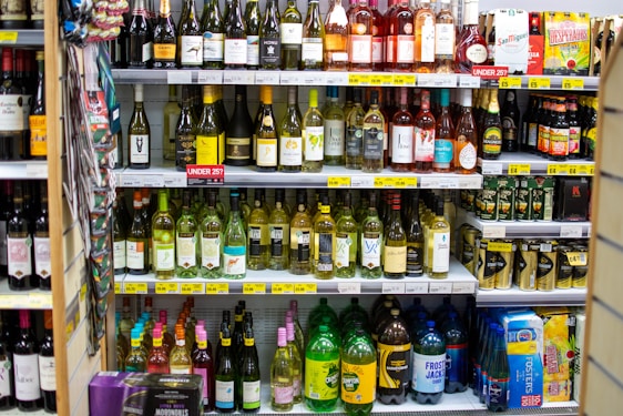 A supermarket shelf filled with various bottles of wine, beer, and cider. The wines are organized by type and brand on the upper shelves, with price labels displayed underneath. On the lower shelves, larger bottles and cans of alcoholic beverages are arranged. The layout is colorful with a mix of glass and plastic packaging.