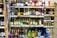 A supermarket shelf filled with various bottles of wine, beer, and cider. The wines are organized by type and brand on the upper shelves, with price labels displayed underneath. On the lower shelves, larger bottles and cans of alcoholic beverages are arranged. The layout is colorful with a mix of glass and plastic packaging.