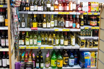 A supermarket shelf filled with various bottles of wine, beer, and cider. The wines are organized by type and brand on the upper shelves, with price labels displayed underneath. On the lower shelves, larger bottles and cans of alcoholic beverages are arranged. The layout is colorful with a mix of glass and plastic packaging.
