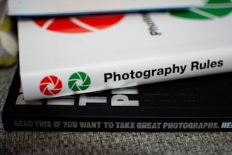 A stack of books with visible spines. The top book is titled 'Photography Rules' with red and green shutter icons, while another book beneath reads 'READ THIS IF YOU WANT TO TAKE GREAT PHOTOGRAPHS.' The surface has a textured fabric appearance.