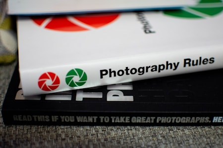 A stack of books with visible spines. The top book is titled 'Photography Rules' with red and green shutter icons, while another book beneath reads 'READ THIS IF YOU WANT TO TAKE GREAT PHOTOGRAPHS.' The surface has a textured fabric appearance.