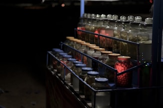 A dimly lit display of jars filled with various preserved food items. The jars are arranged in tiers on metal shelves and are sealed with different colored lids. Some jars are illuminated with red light, creating a warm contrast against the dark background.