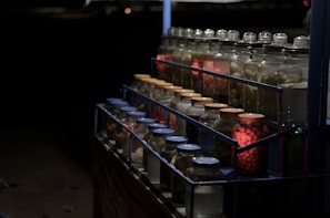Various fermentation starters and koji cultures displayed in small glass jars on a wooden shelf, illuminated by soft natural light.