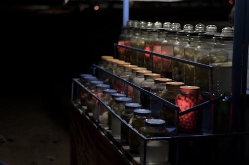 A dimly lit display of jars filled with various preserved food items. The jars are arranged in tiers on metal shelves and are sealed with different colored lids. Some jars are illuminated with red light, creating a warm contrast against the dark background.
