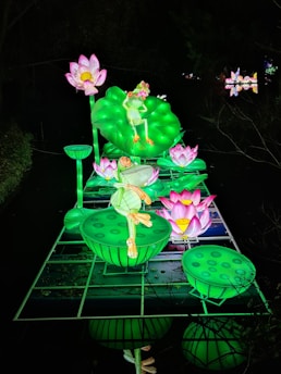 A vibrant, illuminated display features a large green frog poised on a lily pad surrounded by pink and white lotus flowers. The glowing installation is set against a dark background, highlighting the contrast between the bright colors and the shadows.