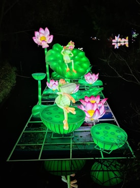 A vibrant, illuminated display features a large green frog poised on a lily pad surrounded by pink and white lotus flowers. The glowing installation is set against a dark background, highlighting the contrast between the bright colors and the shadows.