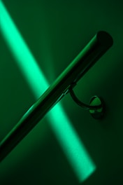 A polished metal handrail is mounted on a wall, with vibrant green lighting casting a bold shadow across the surface.