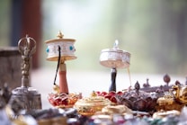 A close-up view of several ornate objects, including prayer wheels and decorative bowls, crafted with intricate metalwork and adorned with jewels and vibrant colors. The background is softly blurred, focusing attention on the artistic details of the items.