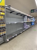 An aisle in a supermarket is shown with mostly empty shelves, except for a few remaining products. There are large bottles of Lipton iced tea on the left and some stacks of toilet paper with purple packaging on the right. The aisle is labeled with a sign for toilet rolls, tissues, and paper towels.