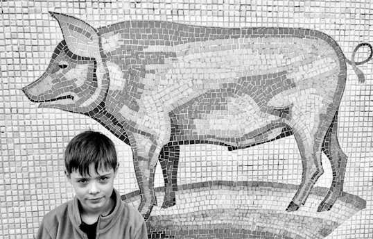 A young boy is standing in front of a large mosaic mural depicting a pig. The mosaic art is composed of numerous square tiles arranged to create the detailed image of the pig.