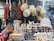 A market stall displays an assortment of wooden kitchen utensils including rolling pins, spoons, and mortar and pestles. Above them, wicker baskets are suspended. To the left, strings of dried vegetables or spices hang as well.