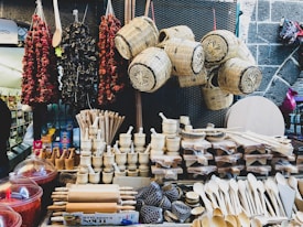 A market stall displays an assortment of wooden kitchen utensils including rolling pins, spoons, and mortar and pestles. Above them, wicker baskets are suspended. To the left, strings of dried vegetables or spices hang as well.