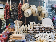 A market stall displays an assortment of wooden kitchen utensils including rolling pins, spoons, and mortar and pestles. Above them, wicker baskets are suspended. To the left, strings of dried vegetables or spices hang as well.