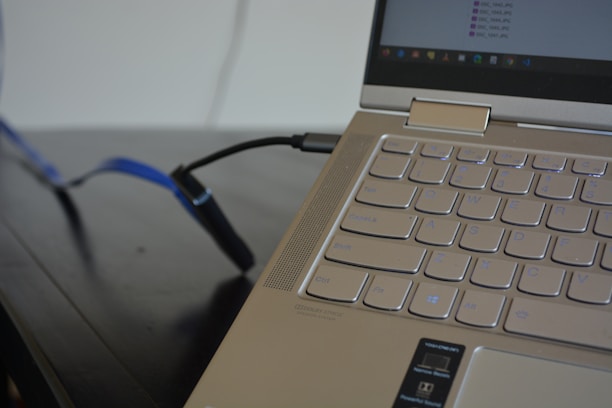 A close-up of a laptop keyboard with a sleek aluminum finish. Several USB ports are visible, and a blue cable is plugged into one of them. The screen shows a file explorer window open with file names partially visible.