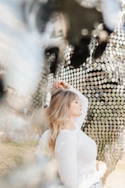 A woman with blonde hair wearing a white long-sleeve top stands in profile with her eyes closed. Her left hand is raised gently over her head while the background features an abstract pattern of reflective discs that create a shimmering effect.