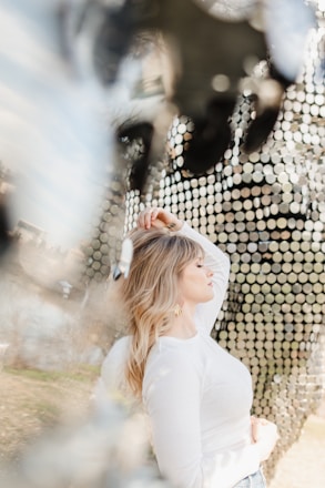 A woman with blonde hair wearing a white long-sleeve top stands in profile with her eyes closed. Her left hand is raised gently over her head while the background features an abstract pattern of reflective discs that create a shimmering effect.