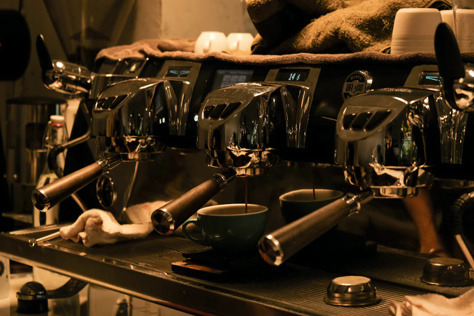 The image shows a commercial coffee machine with three silver nozzles, two on each side and one at the top center. The machine is situated on a countertop, ready to serve customers. A green cup filled with coffee sits atop the machine, indicating that it's in use or about to be used. In front of the machine are several cups and bowls, suggesting that this setup is part of a larger coffee shop or café environment.