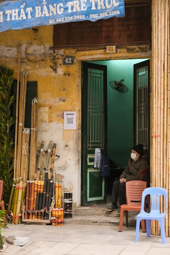 A small shop entrance with a collection of bamboo sticks and crafts displayed outside. A person wearing a mask sits inside the doorway on a red chair. The building has a weathered yellow facade with a blue sign overhead. There are two empty chairs stacked outside next to the bamboo rods.
