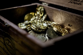 A wooden chest is opened, revealing a pile of shiny gold coins. The coins are illuminated, capturing their metallic luster against the darker interior of the chest. The ambiance suggests an aura of mystery and treasure discovery.