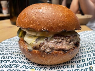 A juicy hamburger with a perfectly toasted bun, a thick beef patty, melted cheese, and pickles. The burger is placed on a branded paper that reads 'BURGER' repeatedly. The background is slightly blurred but suggests a casual dining setting.