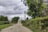A rural dirt road lined with lush green trees and bushes, with a cloudy sky overhead. There are power lines and poles crisscrossing the scene, and a small building and a red barn are visible in the distance.
